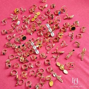 Puede incluir: Una colección de anillos dorados con diversos diseños, incluyendo formas florales, espirales y geométricas. Algunos anillos presentan detalles en negro o conchas blancas. Los anillos están dispuestos sobre una superficie rosa, mostrando una variedad de estilos.