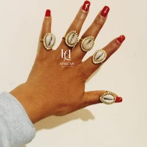 May include: A hand displaying several gold-toned rings, each featuring a white seashell center. The rings are set against a light background. The nails are painted red. The image includes the text "AFRICAN COLLECTION".