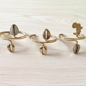 May include: Five gold-toned arm cuffs with unique designs. Each cuff features a curved band with decorative elements, including cowrie shells and an African continent silhouette. The cuffs are displayed on a light-colored wooden surface.
