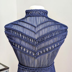 May include: A dark blue macrame garment featuring a high neck collar, intricate chevron-patterned knotting across the shoulders and chest, and a braided waist belt, with long vertical cord fringe extending downward from the waistline.