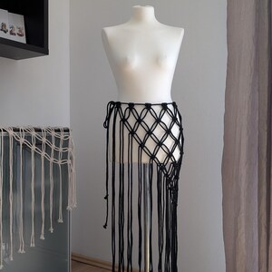Op de afbeelding: Zwarte macramé rok op een mannequin. De rok heeft een ruitvormig patroon en lange franjes. De mannequin staat op een metalen standaard. De achtergrond omvat een muur, een kast en een gordijn.