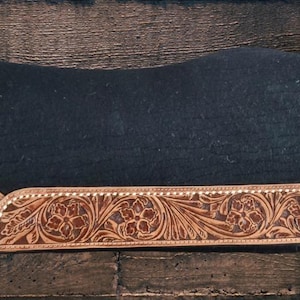 May include: A black saddle pad with a tooled leather border. The border features a floral design in shades of brown and is stitched with a light-colored thread. The pad is rectangular and rests on a dark wood surface.