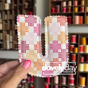May include: A fabric letter U featuring a white background with a repeating grid pattern of pink, orange, and yellow squares. The edges of the letter are scalloped with white stitching. The text "dovely day CREATIVE" is printed in white.