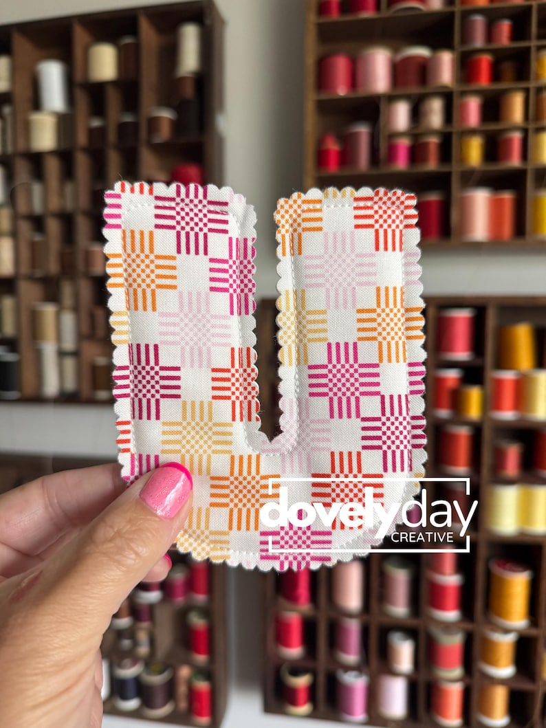 May include: A fabric letter U featuring a white background with a repeating grid pattern of pink, orange, and yellow squares. The edges of the letter are scalloped with white stitching. The text "dovely day CREATIVE" is printed in white.