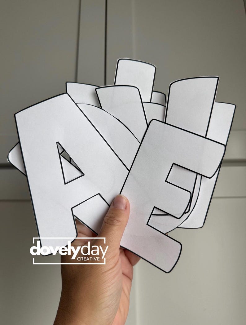 May include: A stack of white paper letters with bold black outlines, featuring the letters A and E prominently in the front. The letters are cut out in a block font style. The logo for dovelyday CREATIVE is printed in white on the bottom left.