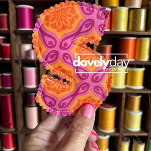 May include: A fabric letter S with a scalloped edge, featuring a vibrant orange and pink floral pattern. The letter is stitched with white thread and displays the white text "dovelyday CREATIVE" centered across the middle of the design.