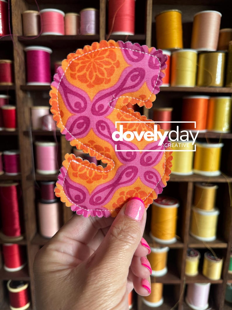 May include: A fabric letter S with a scalloped edge, featuring a vibrant orange and pink floral pattern. The letter is stitched with white thread and displays the white text "dovelyday CREATIVE" centered across the middle of the design.