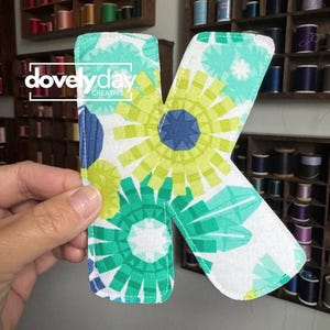 May include: A fabric letter K featuring a white background with a repeating pattern of stylized floral shapes in shades of lime green, teal, and dark blue. The letter has stitched edges and displays the white text dovelyday CREATIVE.