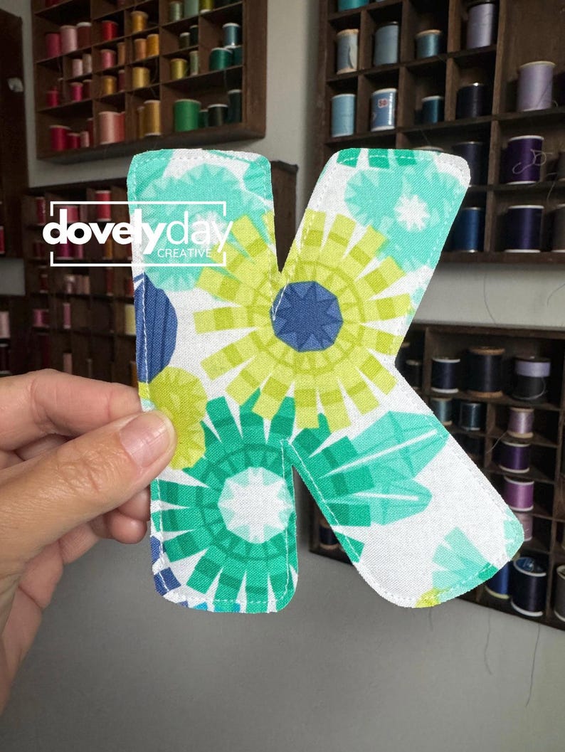 May include: A fabric letter K featuring a white background with a repeating pattern of stylized floral shapes in shades of lime green, teal, and dark blue. The letter has stitched edges and displays the white text dovelyday CREATIVE.