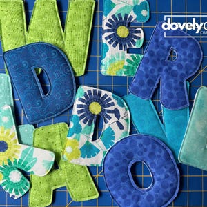 May include: A collection of fabric letters spelling out words, featuring various patterns in shades of blue, green, and teal. The letters include floral prints, polka dots, and swirls. The logo for dovelyday creative is visible in the top right corner.