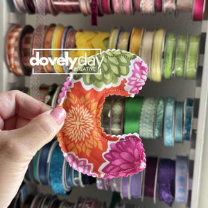 May include: A fabric letter C featuring a vibrant floral pattern in shades of orange, green, and pink. The letter has a scalloped edge with visible stitching and is held against a background of various spools of ribbon organized on a rack.