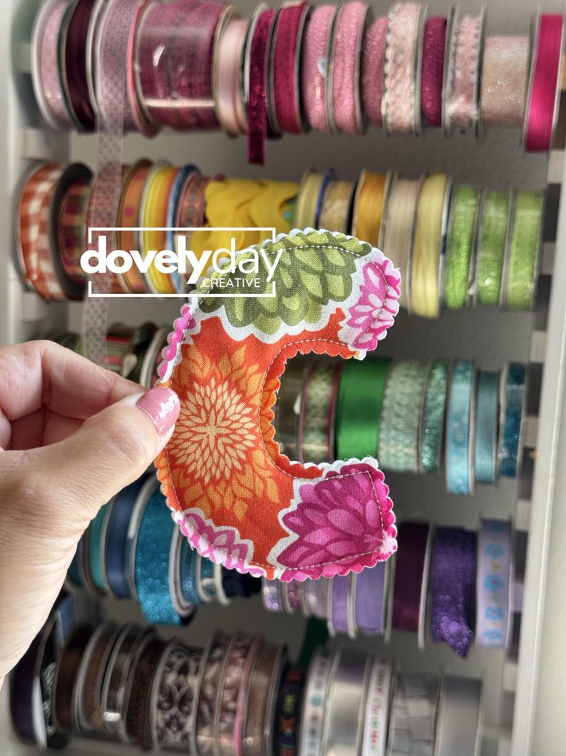 May include: A fabric letter C featuring a vibrant floral pattern in shades of orange, green, and pink. The letter has a scalloped edge with visible stitching and is held against a background of various spools of ribbon organized on a rack.