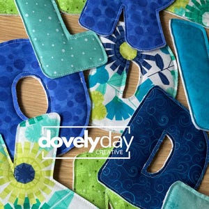 May include: A collection of fabric letters in various shades of blue and green, featuring patterns like polka dots, floral prints, and swirls. The letters are stitched with white thread and include the text "dovelyday CREATIVE" overlaid in white.