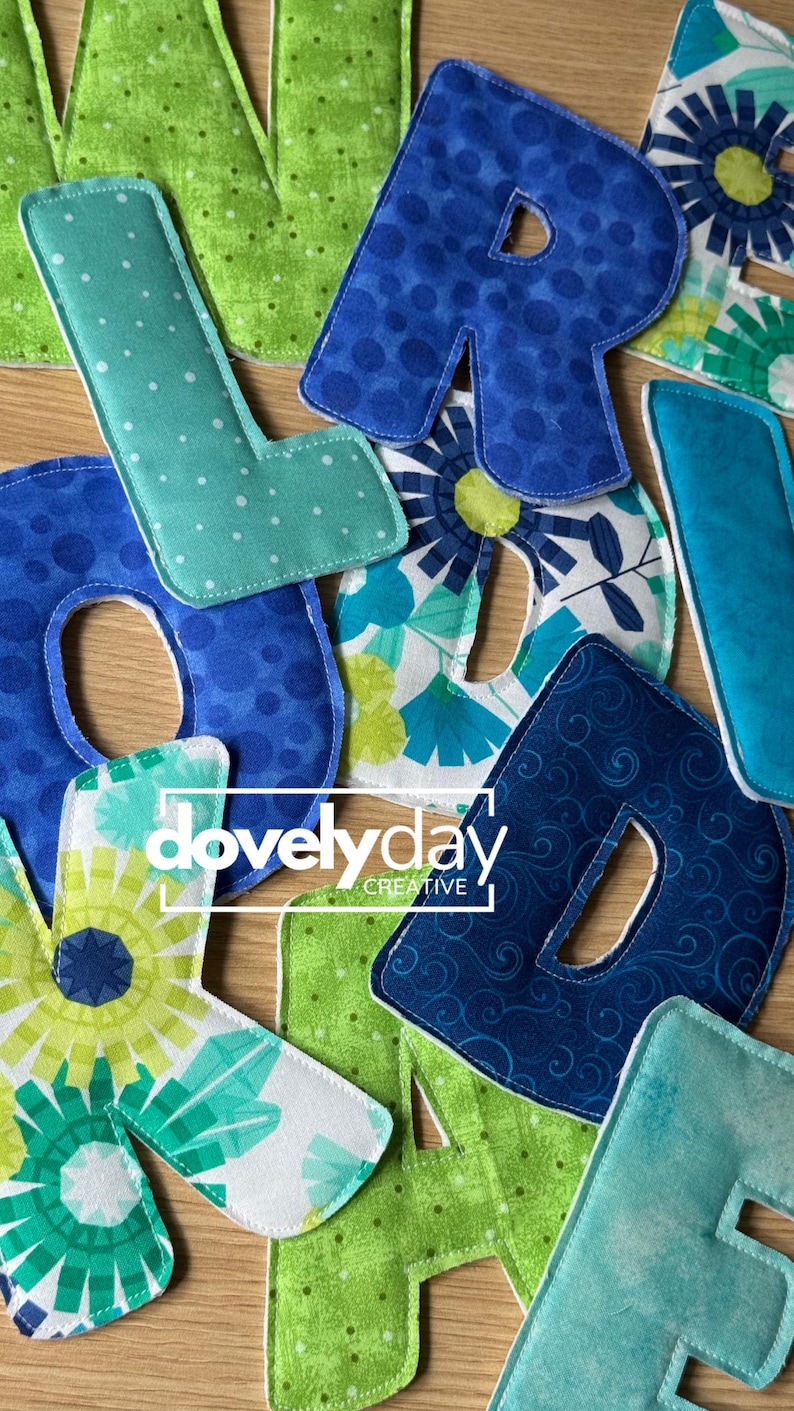 May include: A collection of fabric letters in various shades of blue and green, featuring patterns like polka dots, floral prints, and swirls. The letters are stitched with white thread and include the text "dovelyday CREATIVE" overlaid in white.