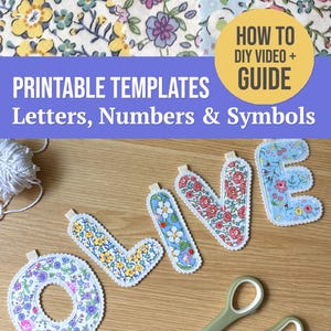 May include: A set of fabric letters spelling LOVE, each featuring distinct floral patterns in yellow, blue, pink, and green. The letters have white scalloped edges and small fabric loops. A pair of green scissors lies nearby on a wooden surface.