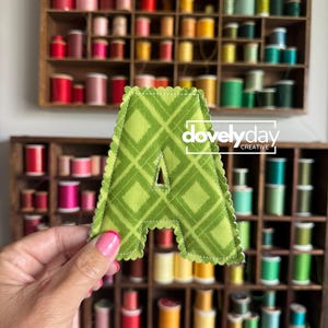 May include: A fabric letter A with a green plaid pattern and scalloped edges. The letter features white stitching along the border and a small triangular cutout in the center. The text "dovelyday CREATIVE" is printed in white over the letter.
