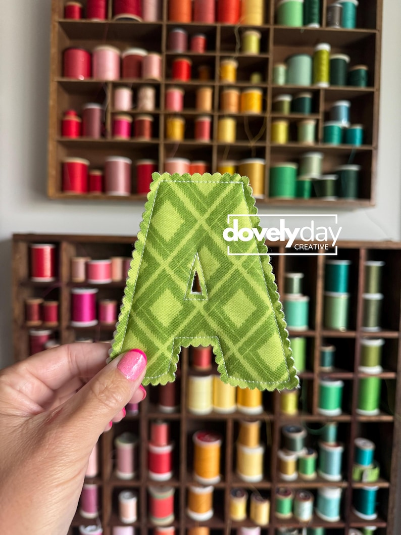 May include: A fabric letter A with a green plaid pattern and scalloped edges. The letter features white stitching along the border and a small triangular cutout in the center. The text "dovelyday CREATIVE" is printed in white over the letter.