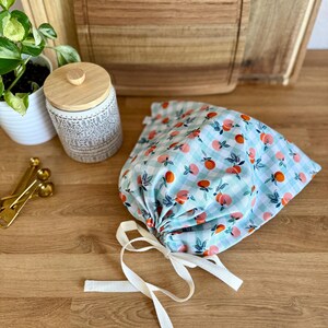 May include: A light blue and white gingham drawstring bag with a pattern of orange and pink peaches and green leaves. The bag is tied with a white ribbon. A wooden cutting board and a decorative jar are in the background.