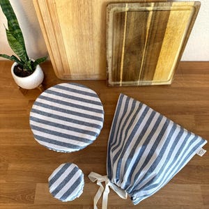 Op de afbeelding: Een verzameling keukenartikelen, waaronder houten snijplanken en gestreepte stoffen voedselhoezen. De hoezen zijn wit met blauwe strepen en zijn er in verschillende maten, waaronder een ronde hoes en een zak met trekkoord. De snijplanken zijn gemaakt van licht en donker hout.