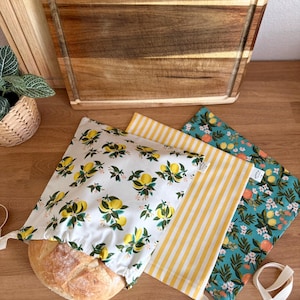 May include: A loaf of bread partially inside a white bread bag with a lemon and floral pattern. Two other bread bags are visible: one with yellow stripes and another with a teal floral pattern. A wooden cutting board is in the background.