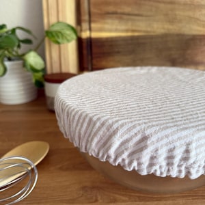 Könnte beinhalten: Eine klare Glasschüssel, bedeckt mit einer beige-weiß gestreiften Stoffschüsselhaube. Ein Holzlöffel und ein Schneebesen befinden sich im Vordergrund. Schneidebretter aus Holz und eine kleine Topfpflanze sind im Hintergrund.