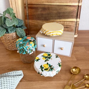 Op de afbeelding: Herbruikbare potdeksels met verschillende patronen. Eén heeft gele en witte strepen, een andere heeft een citroen- en bloemenontwerp op wit en een derde heeft een tropisch bloemenpatroon. Op een houten oppervlak.