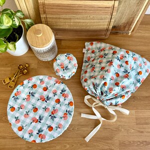 May include: A collection of reusable food covers with a peach and leaf pattern on a light blue and white checkered background. Includes a round cover, a bowl cover, and a drawstring bag. A wooden-topped jar and measuring spoons are also visible.