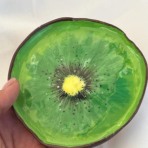 May include: Hand-painted ceramic dish in the shape of a kiwi slice. The dish features a vibrant green interior with a brown rim, a yellow center, and black seed details. The dish is held in a hand.