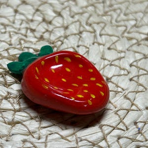May include: A red strawberry-shaped trinket dish with yellow seed details and green leaves. The dish is glossy and sits on a woven surface.