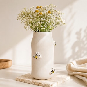 Op de afbeelding: Een cilindrische, gebroken witte keramische vaas met een matte afwerking, versierd met kleine, verhoogde, handgeschilderde hommelmotieven in geel, zwart en wit. De vaas is gevuld met een boeket van witte gipskruid en kleine gele bloemen.