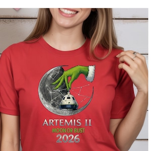 May include: Red t-shirt featuring a graphic of a green hand holding a spacecraft over a crescent moon. The text "ARTEMIS II MOON OR BUST 2026" is printed below the graphic. The design is centered on the shirt.