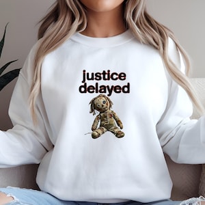 May include: White sweatshirt with the text "justice delayed" in black and red. A distressed doll graphic is centered below the text. The doll has a stitched appearance with button eyes and tattered clothing. The sweatshirt is a casual style.