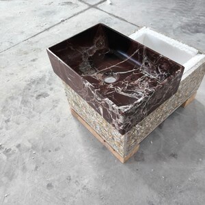 May include: A rectangular, burgundy marble sink with white veining. The sink is set on a multi-colored, textured base. The sink has a single drain hole and is ready for installation. The overall design is modern and luxurious.