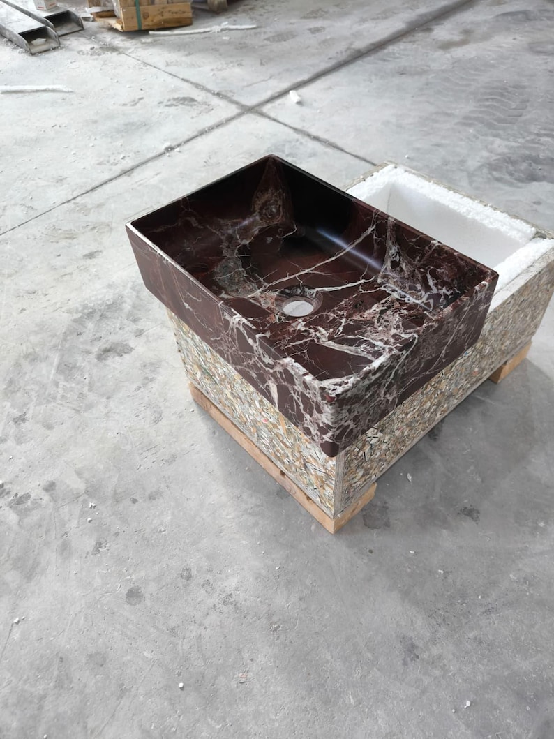 May include: A rectangular, burgundy marble sink with white veining. The sink is set on a multi-colored, textured base. The sink has a single drain hole and is ready for installation. The overall design is modern and luxurious.