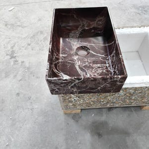 May include: A rectangular bathroom sink made of dark red marble with white veining. The sink has a single drain hole and is set on a multi-colored base. The sink is a modern design.