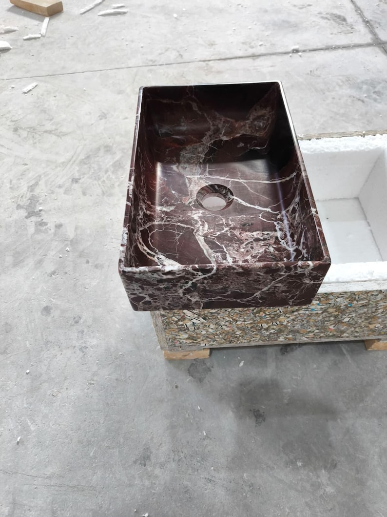 May include: A rectangular bathroom sink made of dark red marble with white veining. The sink has a single drain hole and is set on a multi-colored base. The sink is a modern design.