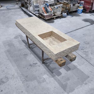 Peut inclure: Un comptoir en pierre de couleur claire avec un évier encastré rectangulaire. Le comptoir est soutenu par un cadre métallique et des poutres en bois. La pierre a une surface rugueuse et texturée.