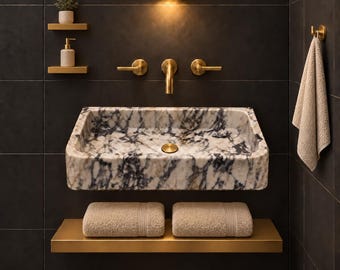 Puede incluir: Un lavabo rectangular de mármol con vetas negras y blancas, con grifo y manijas doradas. Dos estantes pequeños sostienen una planta y un dispensador de jabón, mientras que un estante inferior sostiene dos toallas de color beige.