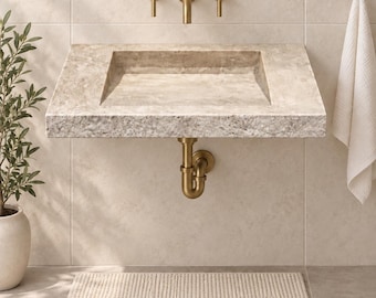 May include: A wall-mounted rectangular stone sink with a textured beige surface, featuring a brass tap and two matching handles. A brass P-trap pipe is visible underneath the basin, which is mounted against a tiled wall with a white towel.