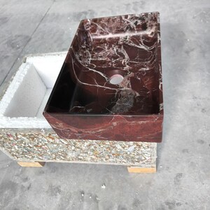 May include: A rectangular bathroom sink with a rich, red marble appearance and white veining. The sink is set on a base made of recycled materials. The sink has a dark interior and a single drain hole.
