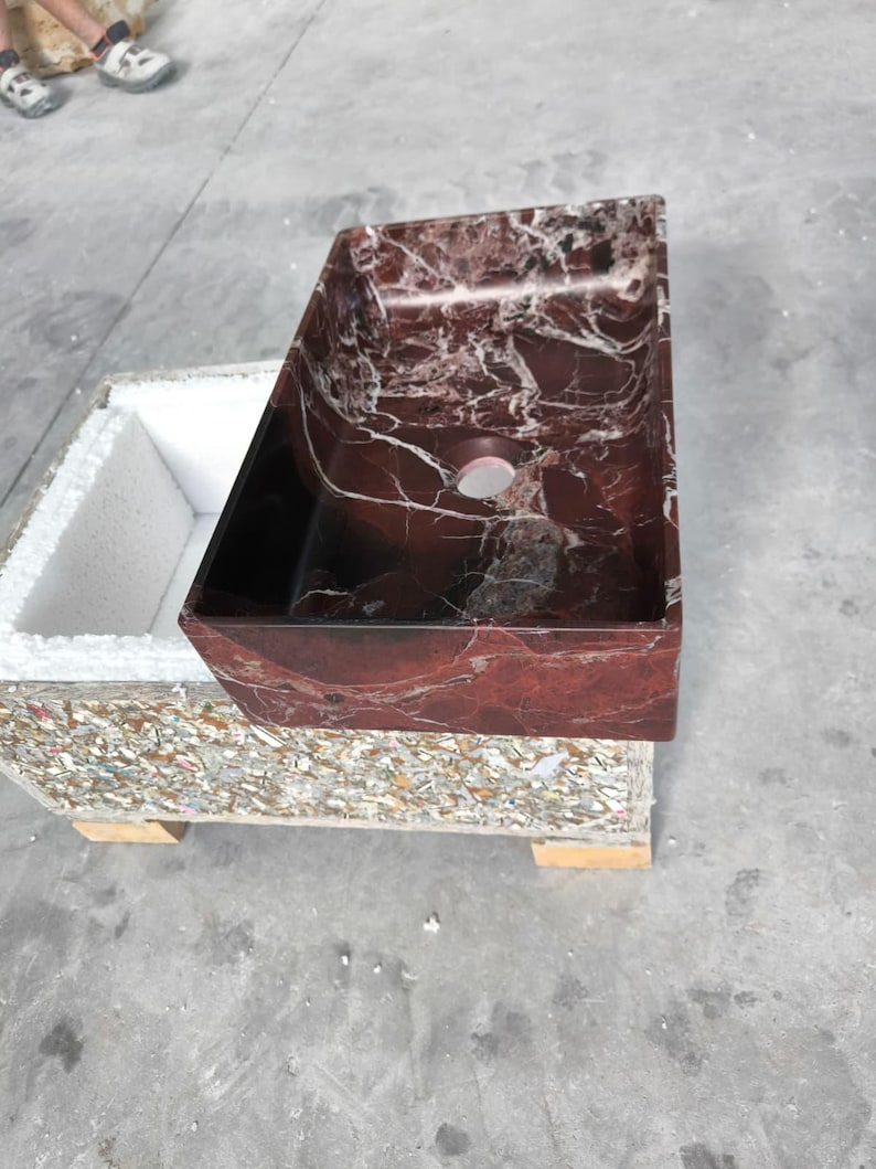 May include: A rectangular bathroom sink with a rich, red marble appearance and white veining. The sink is set on a base made of recycled materials. The sink has a dark interior and a single drain hole.