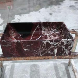 May include: A rectangular sink basin made of burgundy marble with white veining. The basin has a dark interior and is set on a metal frame. The marble has a polished finish, showcasing the natural patterns.