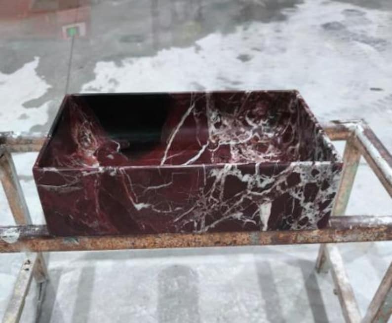 May include: A rectangular sink basin made of burgundy marble with white veining. The basin has a dark interior and is set on a metal frame. The marble has a polished finish, showcasing the natural patterns.