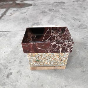 May include: A rectangular, burgundy marble sink with white veining sits atop a composite material base. The sink is displayed on a wooden pallet in a concrete environment. The sink's interior is dark.