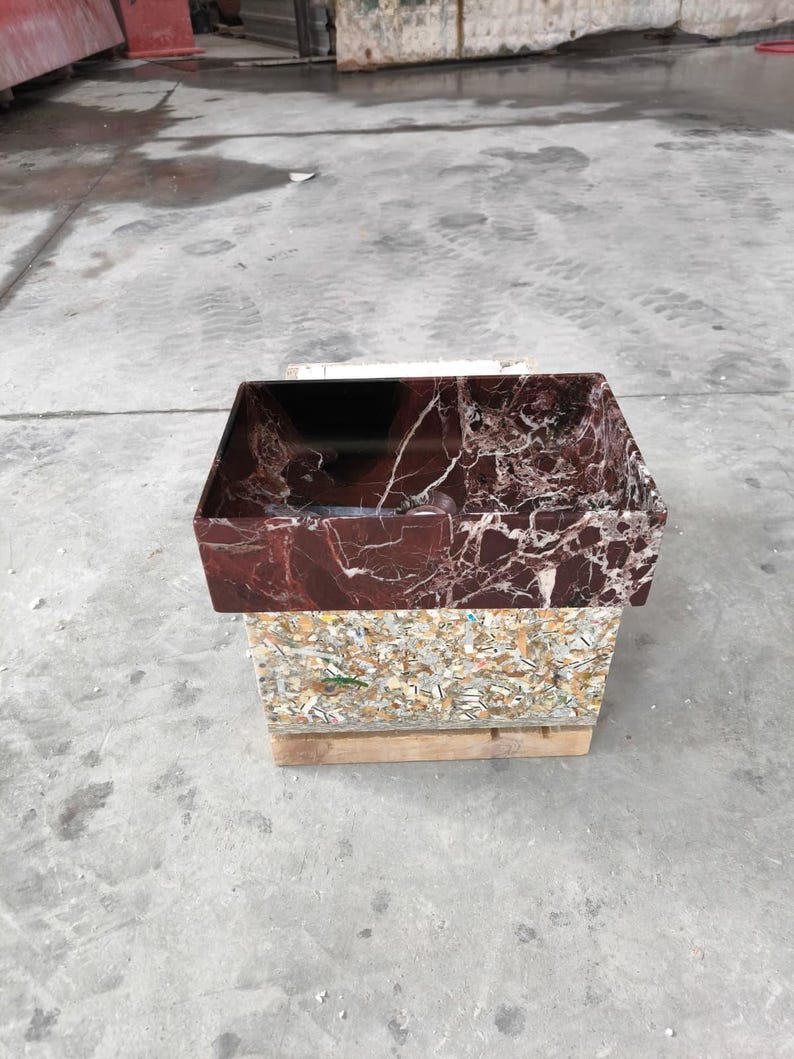 May include: A rectangular, burgundy marble sink with white veining sits atop a composite material base. The sink is displayed on a wooden pallet in a concrete environment. The sink's interior is dark.