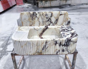 May include: A white marble sink featuring prominent purple and gold veining. The basin is rectangular with a fluted exterior design and an integrated backsplash. The stone surface displays natural color variations throughout the structure.