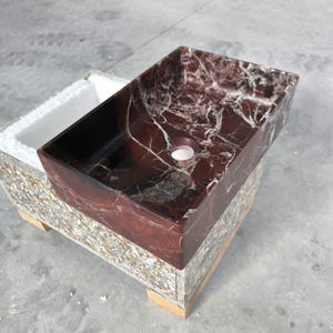 Può includere: Un lavabo rettangolare in marmo rosso con venature bianche. Il lavabo ha un interno scuro e un singolo foro di scarico. Poggia su una base multicolore, probabilmente per la spedizione o l'esposizione.