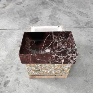 May include: A rectangular, burgundy marble sink with white veining. The sink has a single drain hole and is displayed on a wooden pallet. The sink's rich color and unique pattern make it a striking bathroom fixture.