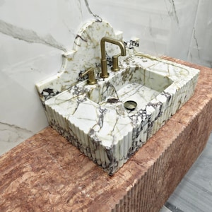 Op de afbeelding: Een marmeren wastafel met gouden kranen. De wastafel is wit met grijze en bruine aderen en een geschulpte rand. De kraan en hendels zijn goudkleurig. De wastafel is gemonteerd op een roodbruine, getextureerde basis.