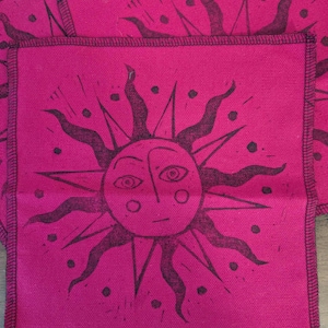 May include: Bright pink square fabric featuring a black sun face design. The sun has a face with eyes, a nose, and a mouth, surrounded by rays and dots. The edges are stitched with black thread.